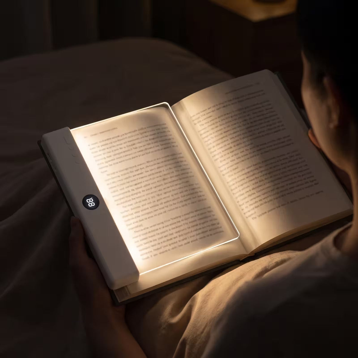 A person holding an open book in a dimly lit room, with a built-in LED light illuminating the pages. The book has a white cover and a small circular control panel on the left edge.