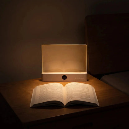 A rectangular LED night light glowing warmly on a wooden bedside table, with an open book placed in front of it in a dimly lit bedroom.