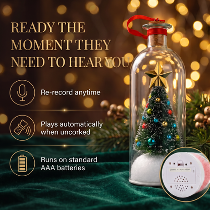A clear glass bottle holiday ornament with a miniature Christmas tree inside, topped with a gold star and a red hanging loop. The bottle sits on a dark green velvet surface with festive pine branches and warm golden bokeh lights in the background.